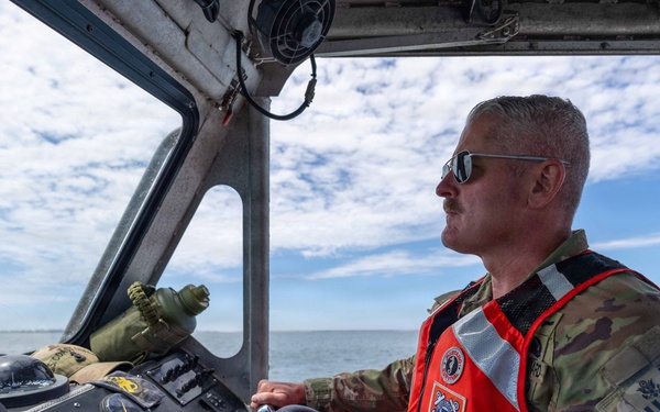A military or Coast Guard personnel member is piloting a patrol boat on open water. The individual is wearing camouflage fatigues and an orange Coast Guard-marked life vest, operating the vessel from a helm console equipped with multiple navigation instruments. The scene appears to be a routine maritime patrol or operational mission under partly cloudy skies.