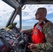 A military or Coast Guard personnel member is piloting a patrol boat on open water. The individual is wearing camouflage fatigues and an orange Coast Guard-marked life vest, operating the vessel from a helm console equipped with multiple navigation instruments. The scene appears to be a routine maritime patrol or operational mission under partly cloudy skies.