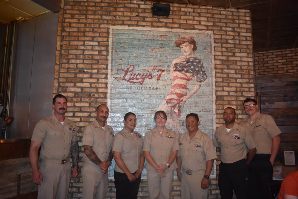 Seven individuals in U.S. Navy tan/khaki uniforms are posing for a group photo inside Lucy's 7 Burger Bar restaurant. They are standing in front of a large brick wall mural featuring a pin-up style woman dressed in an American flag outfit with the restaurant's logo. The setting appears to be a casual dining establishment with rustic brick interior decor.