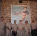 Seven individuals in U.S. Navy tan/khaki uniforms are posing for a group photo inside Lucy's 7 Burger Bar restaurant. They are standing in front of a large brick wall mural featuring a pin-up style woman dressed in an American flag outfit with the restaurant's logo. The setting appears to be a casual dining establishment with rustic brick interior decor.