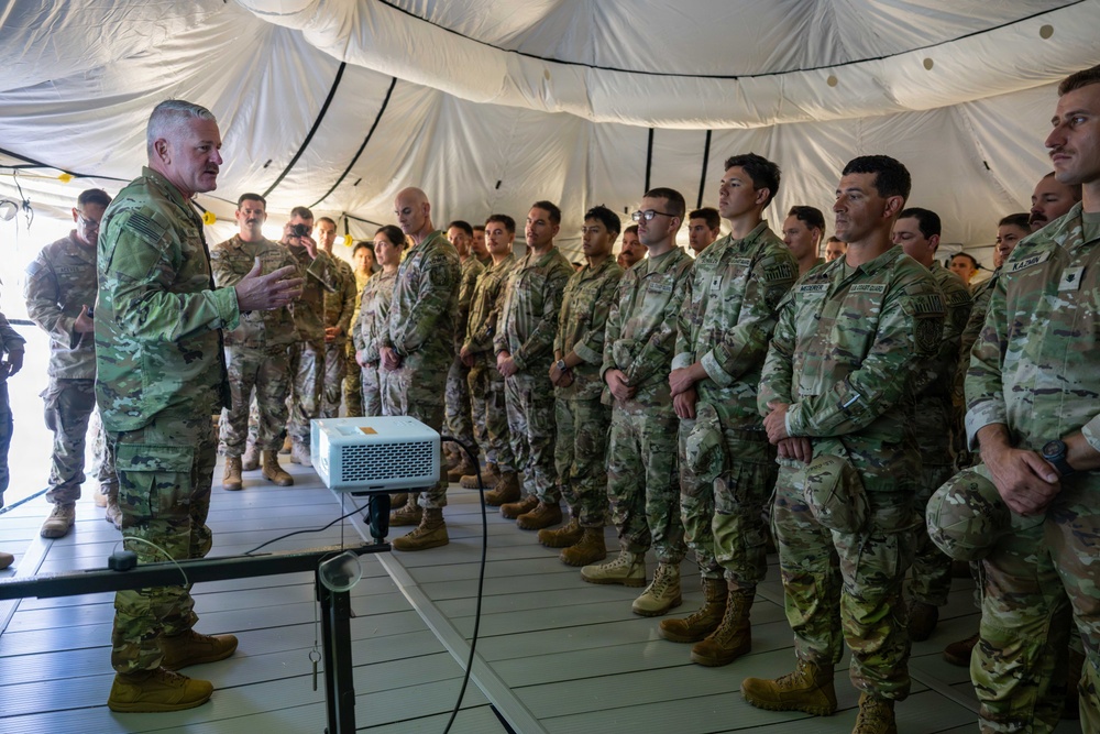 A senior military officer in U.S. Army OCP (Operational Camouflage Pattern) uniform addresses a large group of soldiers inside a military tent or expeditionary shelter. The speaker appears to be conducting a briefing or command address, with a projector visible on a stand in front of him. The assembled soldiers are standing at ease in formation, listening attentively.