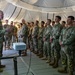 A senior military officer in U.S. Army OCP (Operational Camouflage Pattern) uniform addresses a large group of soldiers inside a military tent or expeditionary shelter. The speaker appears to be conducting a briefing or command address, with a projector visible on a stand in front of him. The assembled soldiers are standing at ease in formation, listening attentively.