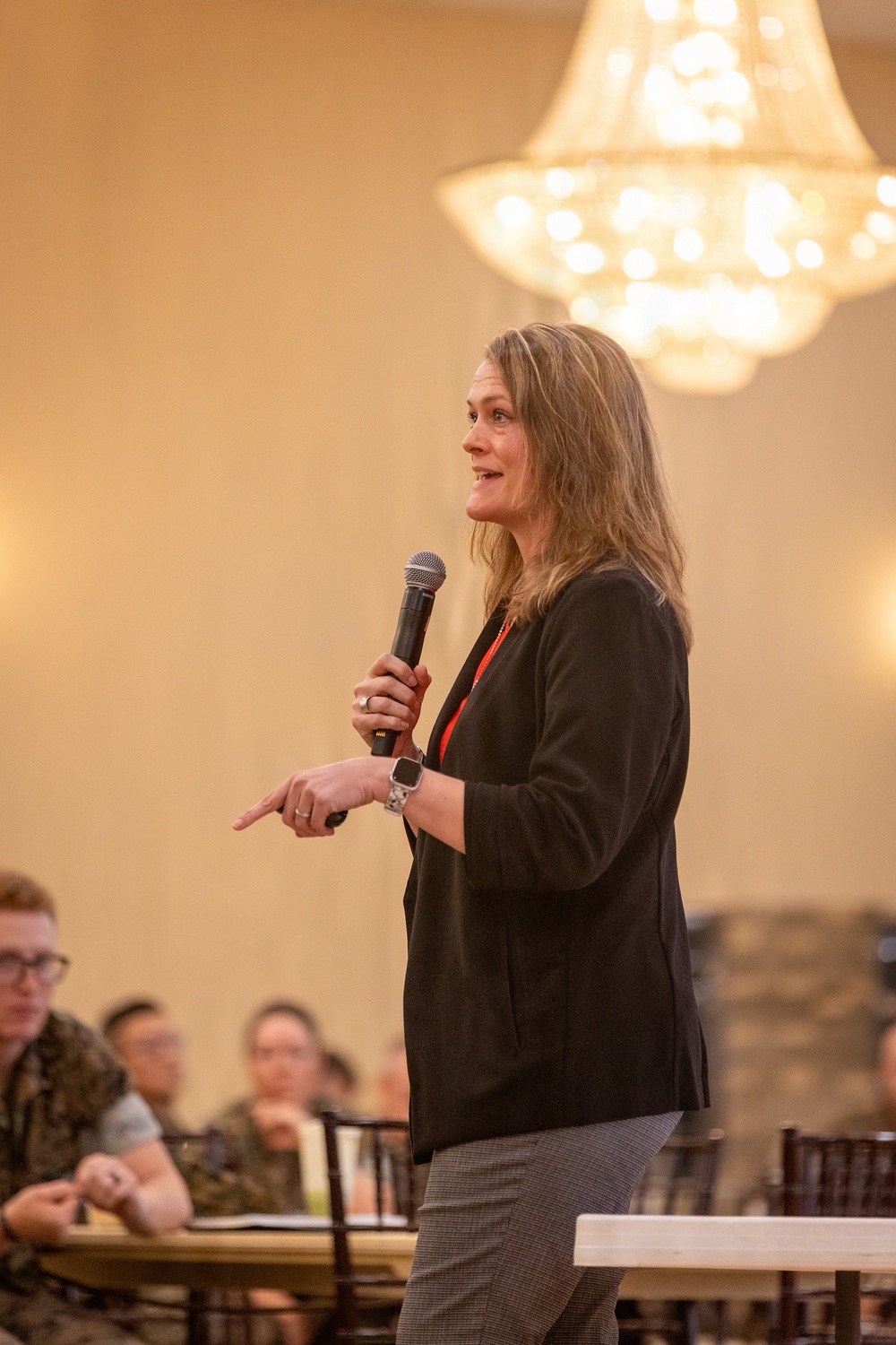 A woman is speaking at a formal event in a banquet hall, holding a handheld microphone and gesturing with her free hand. She is dressed in a black blazer, gray patterned pants, and a red lanyard or necklace, suggesting a professional or conference setting. An audience is seated at round tables in the background beneath a large chandelier.