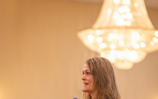 A woman is speaking at a formal event in a banquet hall, holding a handheld microphone and gesturing with her free hand. She is dressed in a black blazer, gray patterned pants, and a red lanyard or necklace, suggesting a professional or conference setting. An audience is seated at round tables in the background beneath a large chandelier.