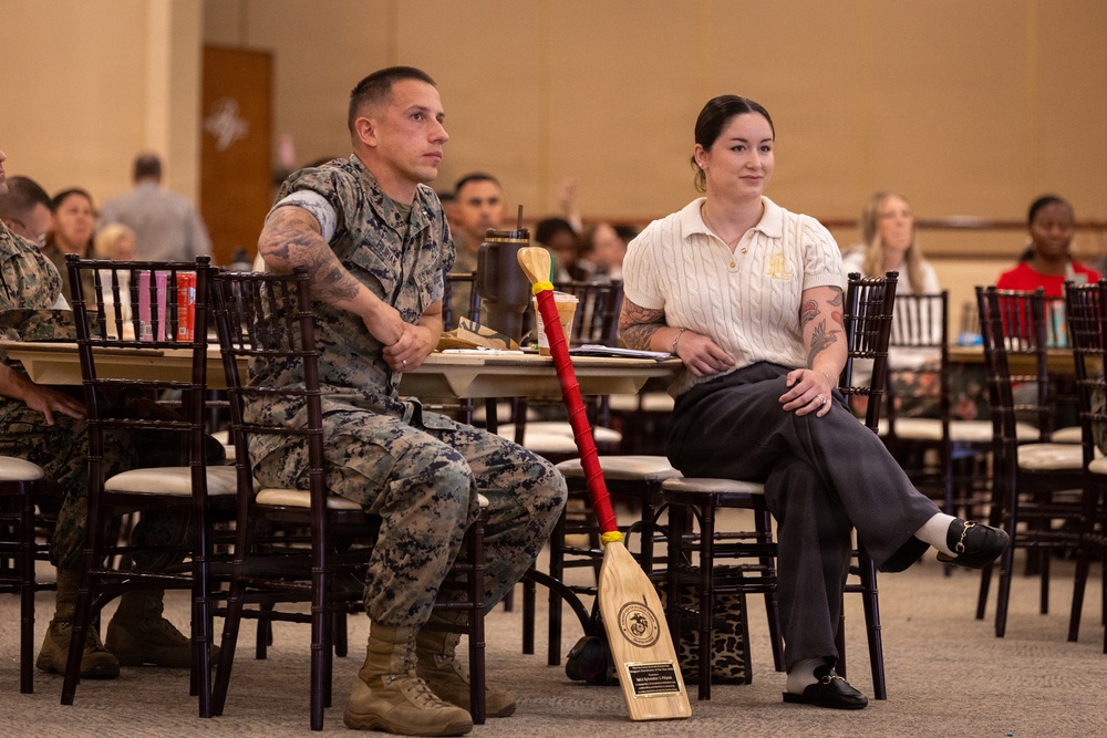 Two individuals are seated at a table in a large meeting or event hall, appearing to attend a formal presentation or ceremony. One person is wearing U.S. Marine Corps camouflage utilities, while the other is in civilian attire. A decorative red-and-wood paddle or oar with a U.S. Marine Corps emblem leans against their table, suggesting an award or recognition event.