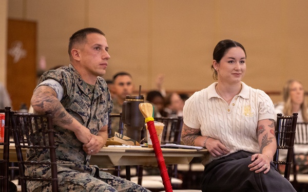Two individuals are seated at a table in a large meeting or event hall, appearing to attend a formal presentation or ceremony. One person is wearing U.S. Marine Corps camouflage utilities, while the other is in civilian attire. A decorative red-and-wood paddle or oar with a U.S. Marine Corps emblem leans against their table, suggesting an award or recognition event.