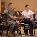 Two individuals are seated at a table in a large meeting or event hall, appearing to attend a formal presentation or ceremony. One person is wearing U.S. Marine Corps camouflage utilities, while the other is in civilian attire. A decorative red-and-wood paddle or oar with a U.S. Marine Corps emblem leans against their table, suggesting an award or recognition event.