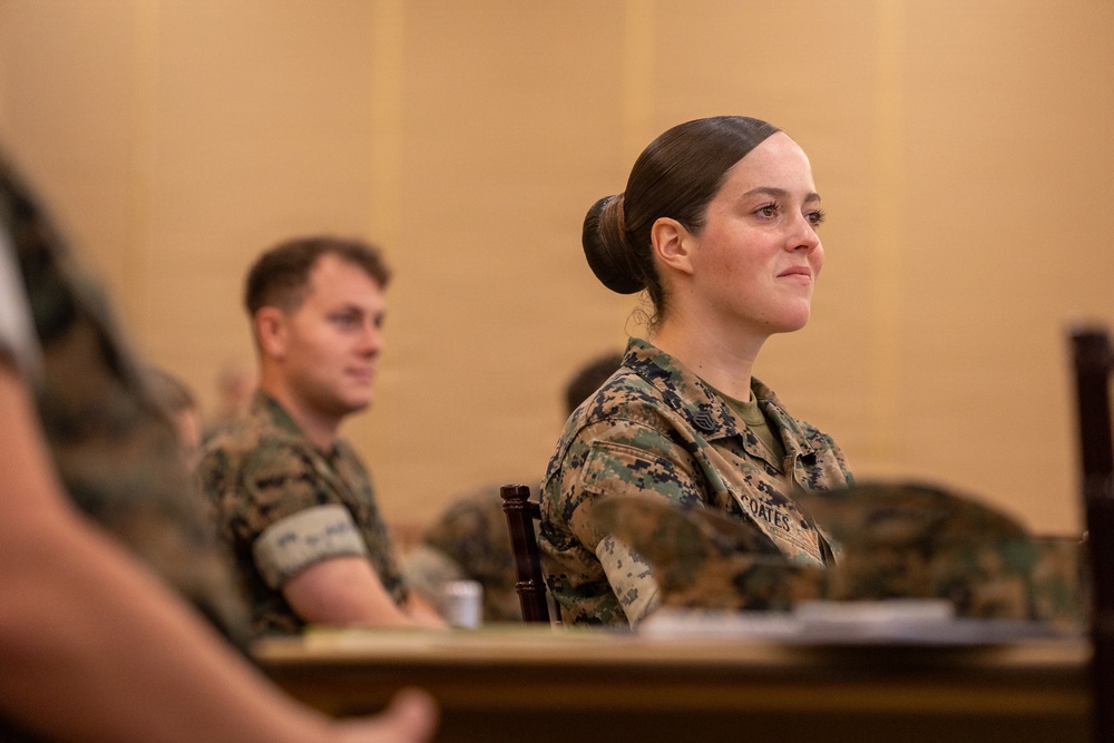 Two U.S. Marines in MARPAT camouflage uniforms are seated at a table, appearing to attend a briefing or classroom instruction session. The foreground Marine, whose name tape reads 'COATES,' is a female with her hair in a regulation bun, attentively facing forward. A male Marine is seated slightly behind and to her left, also in uniform.