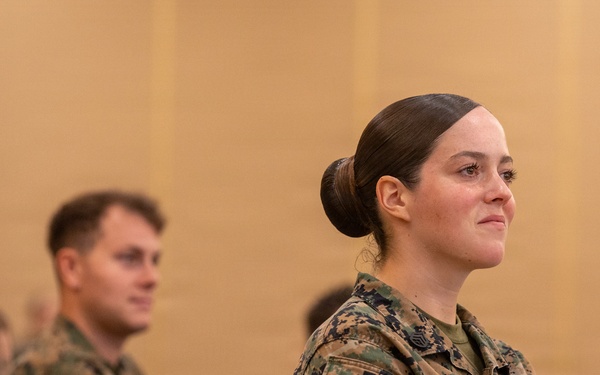 Two U.S. Marines in MARPAT camouflage uniforms are seated at a table, appearing to attend a briefing or classroom instruction session. The foreground Marine, whose name tape reads 'COATES,' is a female with her hair in a regulation bun, attentively facing forward. A male Marine is seated slightly behind and to her left, also in uniform.