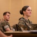Two U.S. Marines in MARPAT camouflage uniforms are seated at a table, appearing to attend a briefing or classroom instruction session. The foreground Marine, whose name tape reads 'COATES,' is a female with her hair in a regulation bun, attentively facing forward. A male Marine is seated slightly behind and to her left, also in uniform.