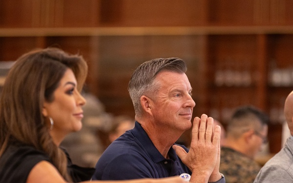 A group of people are seated at round tables in what appears to be a formal banquet or conference hall setting. Three individuals are prominently in focus in the foreground, with military personnel in camouflage uniforms visible in the background. The central male figure has his hands pressed together in an attentive, contemplative posture while observing something off-camera.