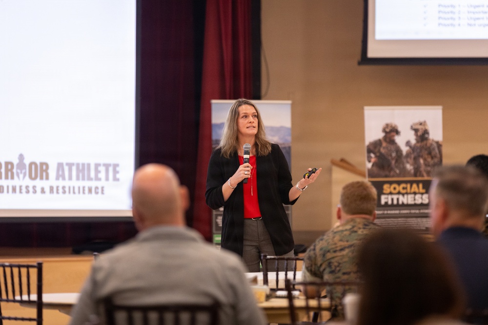 A woman is presenting at a military-themed conference or briefing, holding a microphone and a clicker while addressing an audience. The event appears to be related to the 'Warrior Athlete Readiness &amp; Resilience' program, with visible banners including one labeled 'Social Fitness'. Audience members, including individuals in military camouflage uniforms, are seated at tables facing the speaker.