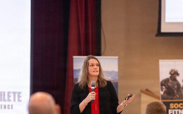 A woman is presenting at a military-themed conference or briefing, holding a microphone and a clicker while addressing an audience. The event appears to be related to the 'Warrior Athlete Readiness &amp; Resilience' program, with visible banners including one labeled 'Social Fitness'. Audience members, including individuals in military camouflage uniforms, are seated at tables facing the speaker.