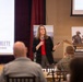 A woman is presenting at a military-themed conference or briefing, holding a microphone and a clicker while addressing an audience. The event appears to be related to the 'Warrior Athlete Readiness &amp; Resilience' program, with visible banners including one labeled 'Social Fitness'. Audience members, including individuals in military camouflage uniforms, are seated at tables facing the speaker.