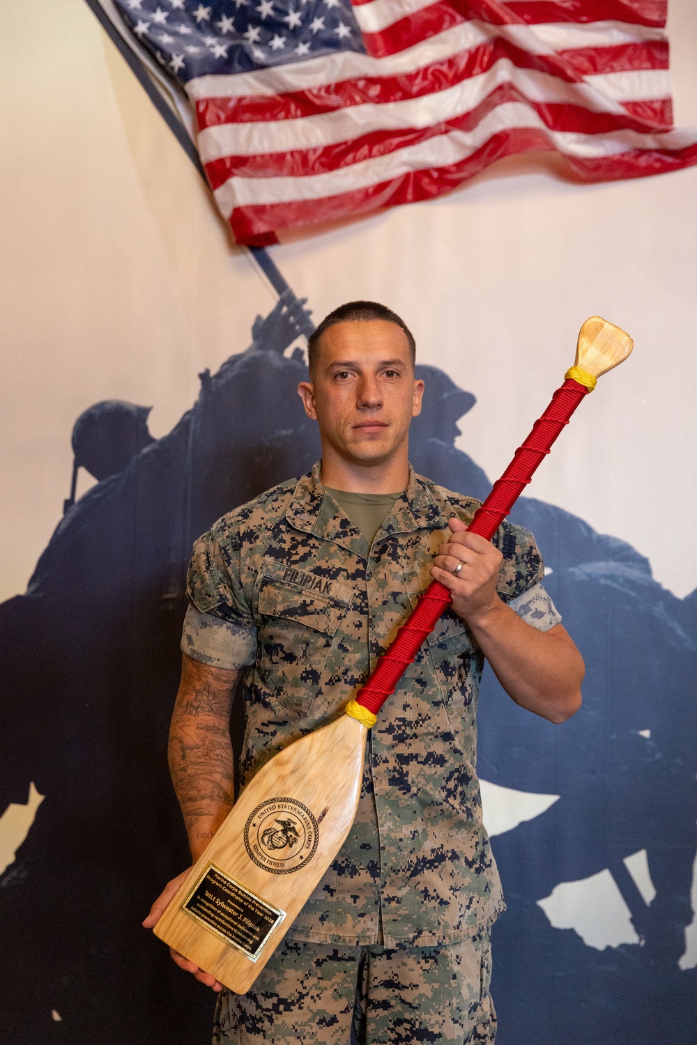 A United States Marine in MARPAT camouflage uniform is posing formally while holding a decorative wooden paddle award with red cord wrapping. The name tape on his uniform reads 'FILIPIAK' and the plaque on the paddle references the United States Marine Corps with 'Semper Fidelis' and the name 'Sylvester J. Filipiak.' He is standing in front of a mural depicting the iconic Iwo Jima flag-raising silhouette, with an American flag displayed above.