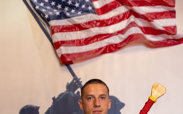 A United States Marine in MARPAT camouflage uniform is posing formally while holding a decorative wooden paddle award with red cord wrapping. The name tape on his uniform reads 'FILIPIAK' and the plaque on the paddle references the United States Marine Corps with 'Semper Fidelis' and the name 'Sylvester J. Filipiak.' He is standing in front of a mural depicting the iconic Iwo Jima flag-raising silhouette, with an American flag displayed above.