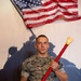 A United States Marine in MARPAT camouflage uniform is posing formally while holding a decorative wooden paddle award with red cord wrapping. The name tape on his uniform reads 'FILIPIAK' and the plaque on the paddle references the United States Marine Corps with 'Semper Fidelis' and the name 'Sylvester J. Filipiak.' He is standing in front of a mural depicting the iconic Iwo Jima flag-raising silhouette, with an American flag displayed above.