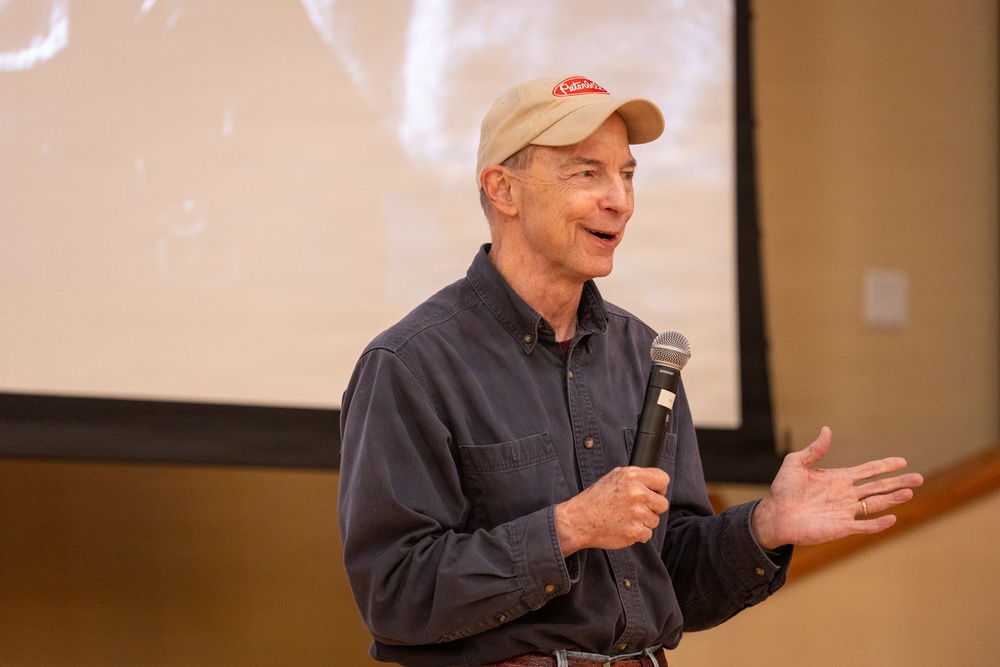 An older man is speaking into a handheld microphone in what appears to be a presentation or lecture setting. He is wearing a beige baseball cap with a red logo and a dark blue button-up shirt. A projection screen is visible in the background, displaying a faded black-and-white image.