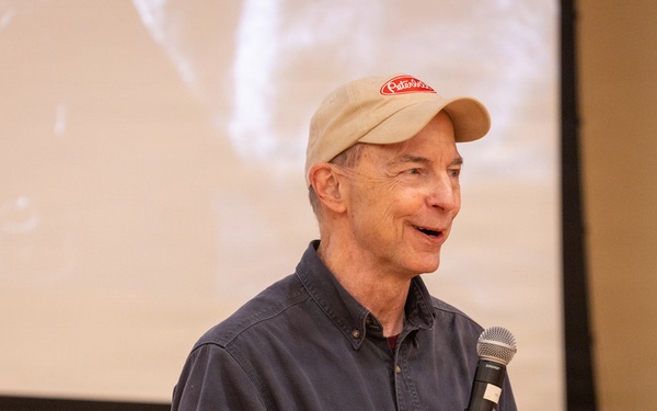 An older man is speaking into a handheld microphone in what appears to be a presentation or lecture setting. He is wearing a beige baseball cap with a red logo and a dark blue button-up shirt. A projection screen is visible in the background, displaying a faded black-and-white image.