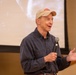An older man is speaking into a handheld microphone in what appears to be a presentation or lecture setting. He is wearing a beige baseball cap with a red logo and a dark blue button-up shirt. A projection screen is visible in the background, displaying a faded black-and-white image.