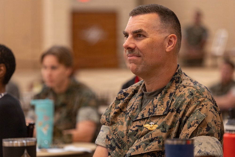 A U.S. Navy officer wearing Marine Corps MARPAT digital camouflage utilities is seated at what appears to be a formal dining or briefing event. The officer displays a name tape reading 'HATYMAN' (or similar) and 'U.S. NAVY,' along with visible gold insignia including what appears to be a naval aviation or command badge. The background shows other attendees seated at tables in a large hall, rendered out of focus.