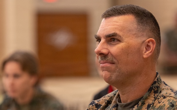 A U.S. Navy officer wearing Marine Corps MARPAT digital camouflage utilities is seated at what appears to be a formal dining or briefing event. The officer displays a name tape reading 'HATYMAN' (or similar) and 'U.S. NAVY,' along with visible gold insignia including what appears to be a naval aviation or command badge. The background shows other attendees seated at tables in a large hall, rendered out of focus.