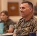 A U.S. Navy officer wearing Marine Corps MARPAT digital camouflage utilities is seated at what appears to be a formal dining or briefing event. The officer displays a name tape reading 'HATYMAN' (or similar) and 'U.S. NAVY,' along with visible gold insignia including what appears to be a naval aviation or command badge. The background shows other attendees seated at tables in a large hall, rendered out of focus.