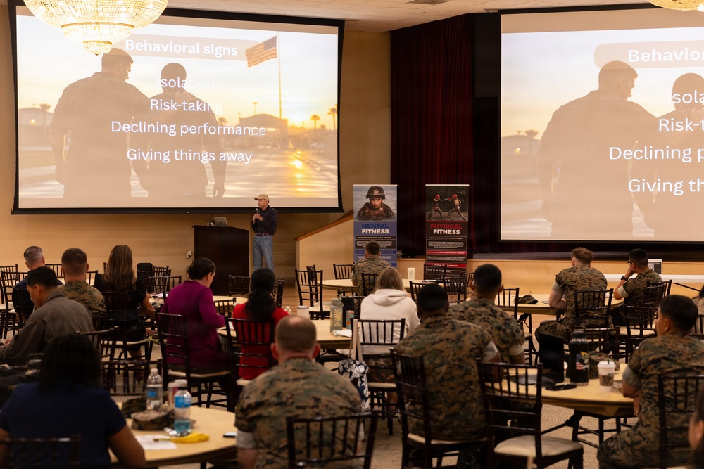 A presenter is delivering a mental health or wellness briefing to a room of military personnel and civilians, with two large projection screens displaying a slide titled 'Behavioral Signs' listing warning indicators such as isolation, risk-taking, declining performance, and giving things away. The audience is seated at round tables in a banquet-style hall, with the majority wearing Marine Corps camouflage uniforms. Two promotional banners labeled 'Mental Fitness' and 'Physical Fitness' are visible between the screens.