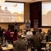 A presenter is delivering a mental health or wellness briefing to a room of military personnel and civilians, with two large projection screens displaying a slide titled 'Behavioral Signs' listing warning indicators such as isolation, risk-taking, declining performance, and giving things away. The audience is seated at round tables in a banquet-style hall, with the majority wearing Marine Corps camouflage uniforms. Two promotional banners labeled 'Mental Fitness' and 'Physical Fitness' are visible between the screens.