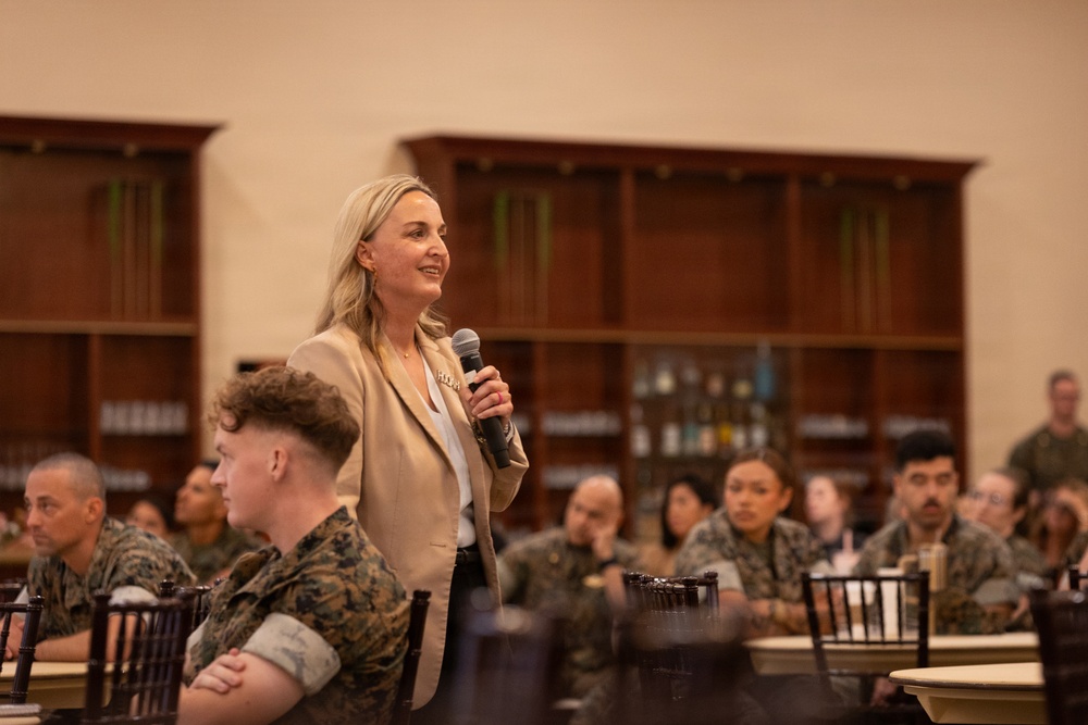 A blonde woman in a tan blazer is speaking into a microphone before an audience of U.S. Marine Corps personnel in camouflage uniforms. The setting appears to be a formal dining hall or event space with dark wood bookshelves in the background. The audience is seated at round tables and appears attentive.