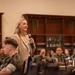 A blonde woman in a tan blazer is speaking into a microphone before an audience of U.S. Marine Corps personnel in camouflage uniforms. The setting appears to be a formal dining hall or event space with dark wood bookshelves in the background. The audience is seated at round tables and appears attentive.