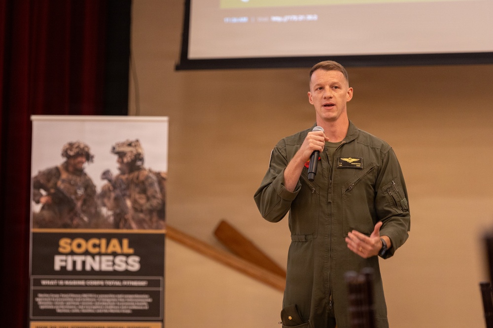 A military officer in an olive green flight suit is speaking at a podium or presentation area, holding a microphone. He appears to be delivering a talk or briefing in front of a projection screen. A banner behind him reads 'Social Fitness' with text referencing 'Marine Corps Total Fitness.'