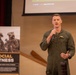 A military officer in an olive green flight suit is speaking at a podium or presentation area, holding a microphone. He appears to be delivering a talk or briefing in front of a projection screen. A banner behind him reads 'Social Fitness' with text referencing 'Marine Corps Total Fitness.'