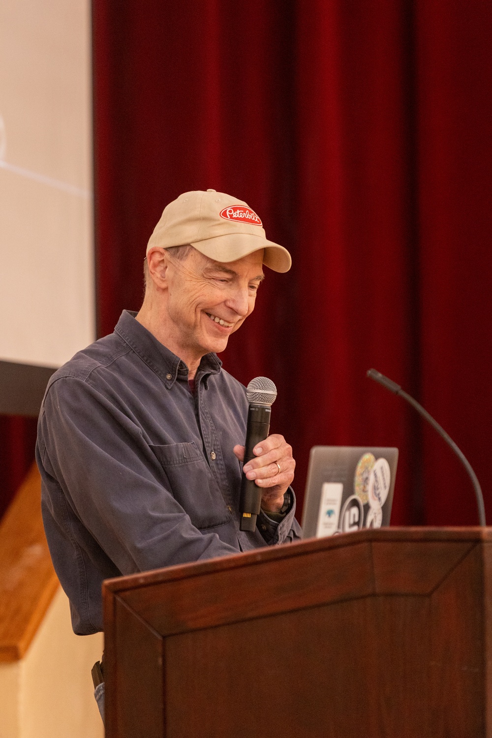 A middle-aged man is speaking at a wooden podium in what appears to be an auditorium or lecture hall. He is smiling and holding a handheld microphone, with a laptop covered in stickers visible on the podium. A red curtain and a projection screen are visible in the background.