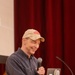 A middle-aged man is speaking at a wooden podium in what appears to be an auditorium or lecture hall. He is smiling and holding a handheld microphone, with a laptop covered in stickers visible on the podium. A red curtain and a projection screen are visible in the background.