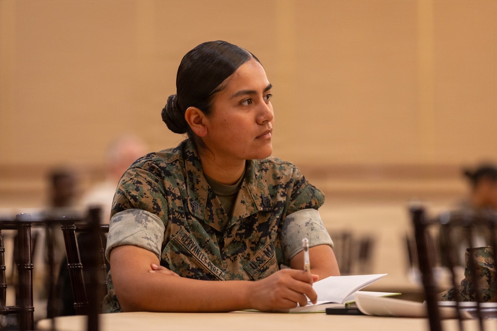A U.S. Marine in MARPAT camouflage uniform is seated at a table in what appears to be a briefing or training session. The Marine, whose name tape reads 'CERVANTES-PEREZ' and wears a rank insignia consistent with a Sergeant, is attentively listening while holding a pen over an open notebook. The setting appears to be a formal indoor venue such as a conference or ballroom facility.