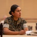 A U.S. Marine in MARPAT camouflage uniform is seated at a table in what appears to be a briefing or training session. The Marine, whose name tape reads 'CERVANTES-PEREZ' and wears a rank insignia consistent with a Sergeant, is attentively listening while holding a pen over an open notebook. The setting appears to be a formal indoor venue such as a conference or ballroom facility.