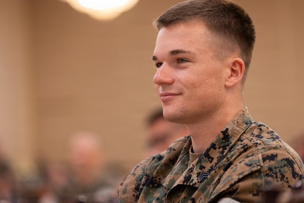 A young male U.S. Marine in MARPAT woodland camouflage uniform is seated in what appears to be a briefing room or auditorium. He is smiling slightly and looking to his left, suggesting he is attentively watching a presentation or speaker. A rank insignia consistent with Sergeant (E-5) is visible on his collar.