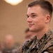 A young male U.S. Marine in MARPAT woodland camouflage uniform is seated in what appears to be a briefing room or auditorium. He is smiling slightly and looking to his left, suggesting he is attentively watching a presentation or speaker. A rank insignia consistent with Sergeant (E-5) is visible on his collar.