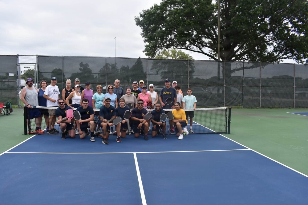A large group of approximately 25-30 people pose together on a blue pickleball court for a group photo. Several participants in the front row are crouching and holding pickleball paddles, wearing matching dark shirts, while others stand behind them. The setting appears to be an outdoor recreational facility with chain-link fencing and windscreen barriers.