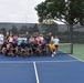 A large group of approximately 25-30 people pose together on a blue pickleball court for a group photo. Several participants in the front row are crouching and holding pickleball paddles, wearing matching dark shirts, while others stand behind them. The setting appears to be an outdoor recreational facility with chain-link fencing and windscreen barriers.