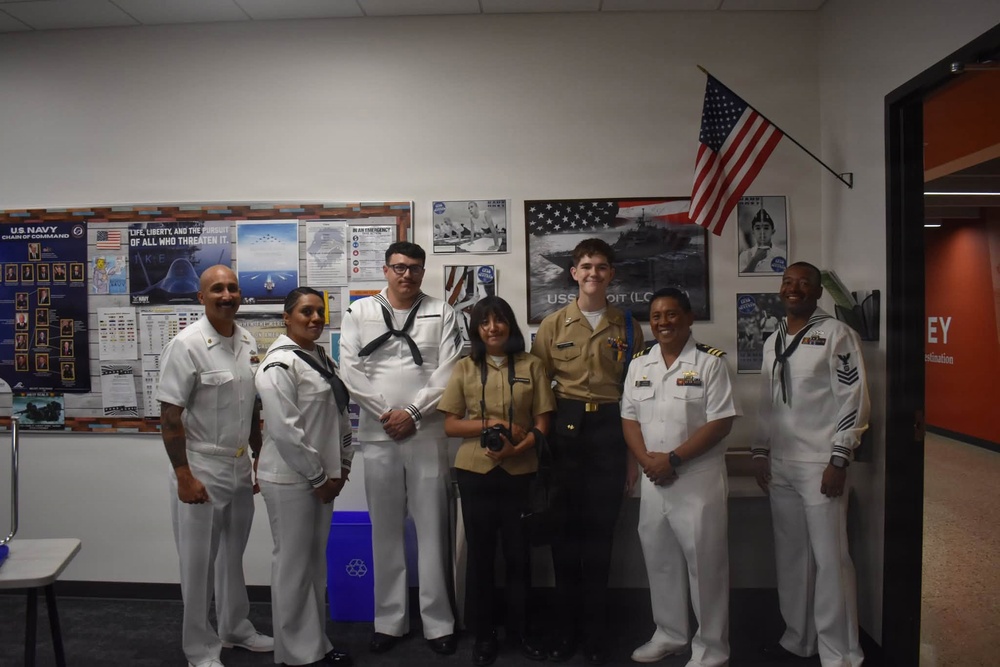 A group of seven individuals pose for a photo in what appears to be a U.S. Navy recruiting or JROTC office. Six of the individuals are wearing U.S. Navy uniforms (white dress uniforms and one in khaki), while one civilian woman stands in the center holding a camera. The room is decorated with U.S. Navy posters, a bulletin board, and an American flag mounted on the wall.