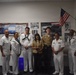 A group of seven individuals pose for a photo in what appears to be a U.S. Navy recruiting or JROTC office. Six of the individuals are wearing U.S. Navy uniforms (white dress uniforms and one in khaki), while one civilian woman stands in the center holding a camera. The room is decorated with U.S. Navy posters, a bulletin board, and an American flag mounted on the wall.