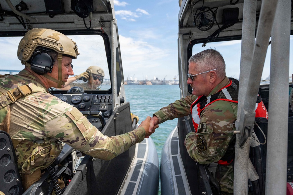 Two military personnel in camouflage uniforms are shaking hands between two patrol boats on open water, with a port or naval facility visible in the background. The individual on the left wears a tactical helmet with communications headset, while the individual on the right wears a red life vest over his uniform and sunglasses. A third person is partially visible in the background operating one of the vessels.