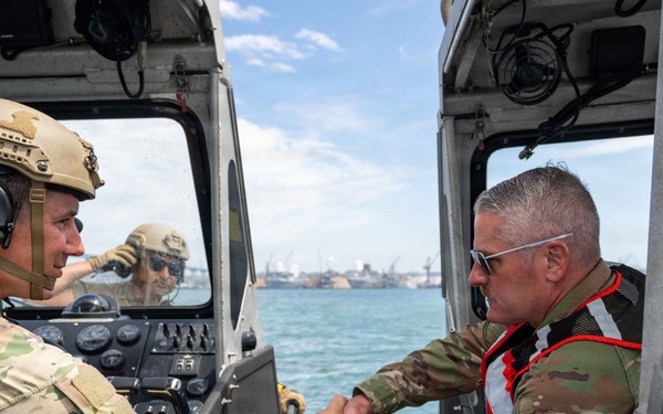 Two military personnel in camouflage uniforms are shaking hands between two patrol boats on open water, with a port or naval facility visible in the background. The individual on the left wears a tactical helmet with communications headset, while the individual on the right wears a red life vest over his uniform and sunglasses. A third person is partially visible in the background operating one of the vessels.