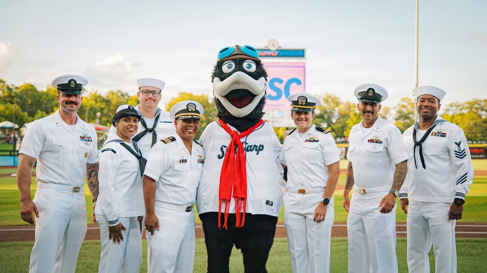 A group of seven U.S. Navy personnel in white dress uniforms pose on a baseball field with a team mascot. The mascot appears to be a bird character wearing a jersey that reads 'Sea Carp' with a red scarf and blue goggles. The setting is a minor league baseball stadium during evening hours.