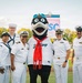 A group of seven U.S. Navy personnel in white dress uniforms pose on a baseball field with a team mascot. The mascot appears to be a bird character wearing a jersey that reads 'Sea Carp' with a red scarf and blue goggles. The setting is a minor league baseball stadium during evening hours.