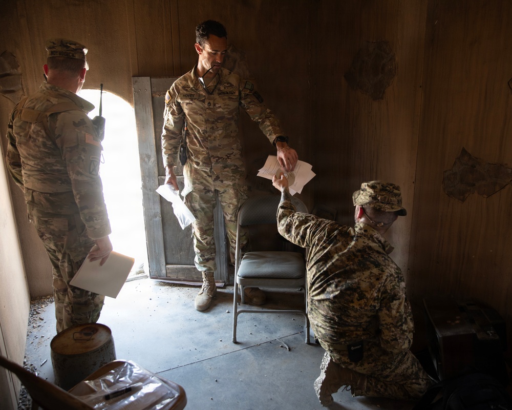 Three military personnel are inside a sparse, worn interior space exchanging documents. Two soldiers in US Army OCP (Operational Camouflage Pattern) uniforms are standing, while a third individual in a different camouflage pattern (possibly a foreign military uniform) is crouching and handing papers upward. The setting appears to be a dilapidated or austere building, possibly a training or operational environment.