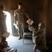 Three military personnel are inside a sparse, worn interior space exchanging documents. Two soldiers in US Army OCP (Operational Camouflage Pattern) uniforms are standing, while a third individual in a different camouflage pattern (possibly a foreign military uniform) is crouching and handing papers upward. The setting appears to be a dilapidated or austere building, possibly a training or operational environment.