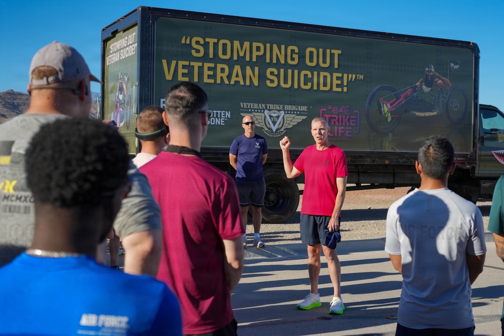 A group of people are gathered outdoors in front of a large truck bearing a 'Stomping Out Veteran Suicide!' billboard from the Veteran Trike Brigade organization. A man in a red shirt appears to be addressing the group with a raised fist, while another man in a blue shirt stands nearby. The setting appears to be a parking area with mountains visible in the background.